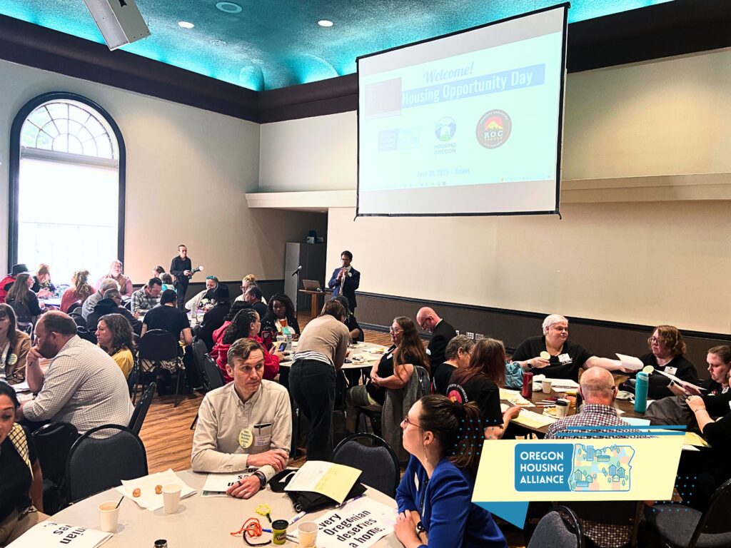 groups of people sitting at tables in a large room with a blue ceiling and a giant window. there's also a projection screen displaying the words "Welcome! Housing Opportunity Day"
