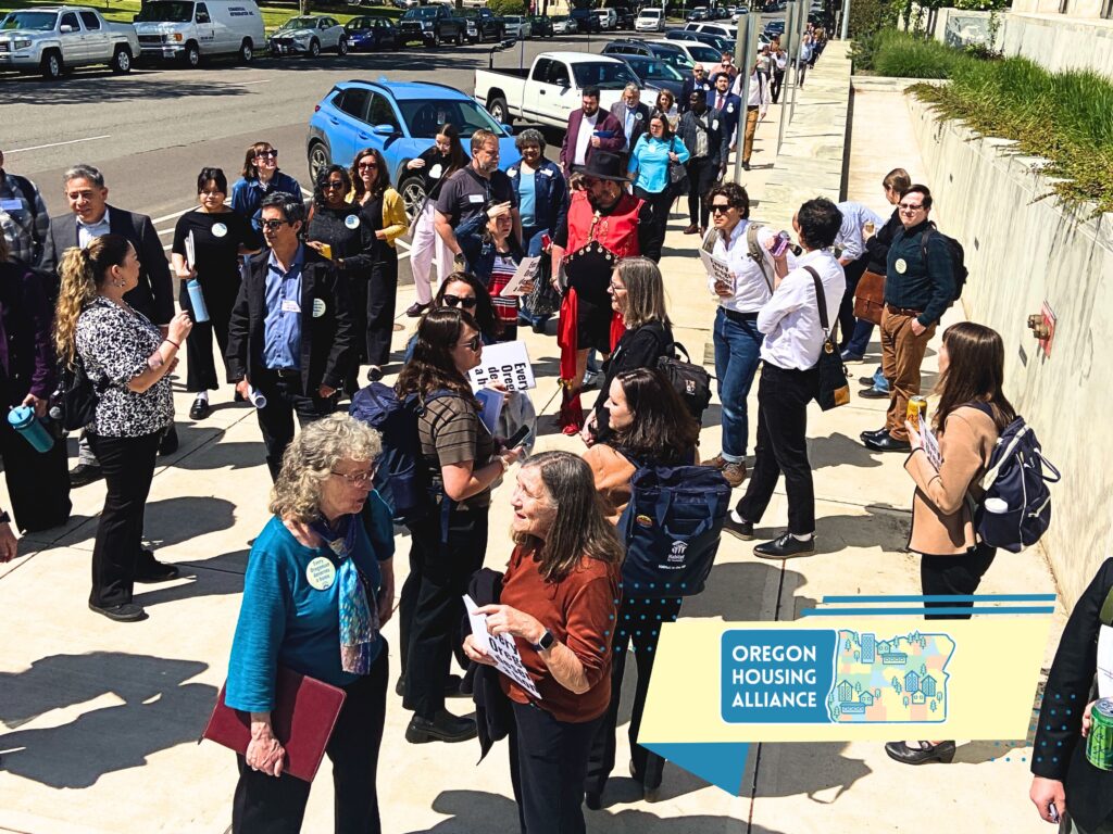 long line of people walking down the street and talking on the sidewalk going back as far as is visible, street is lined with cars parked.
