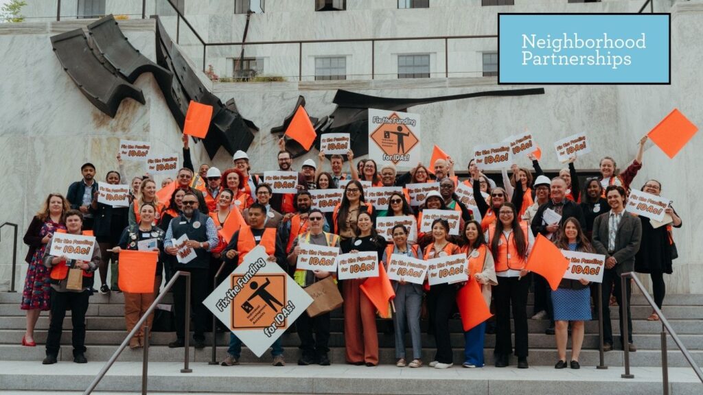 Large group of approximately 75 advocates gathered on the steps of the Oregon State Capitol building, holding bright orange signs reading 'Fix the Funding for IDAs!' Many participants wear orange clothing and hold orange diamond-shaped signs. The diverse crowd includes people of various ages and backgrounds, all smiling and raising their signs enthusiastically. The imposing concrete architecture of the capitol building with tall columns forms the backdrop, with the Neighborhood Partnerships logo visible in the upper right corner.