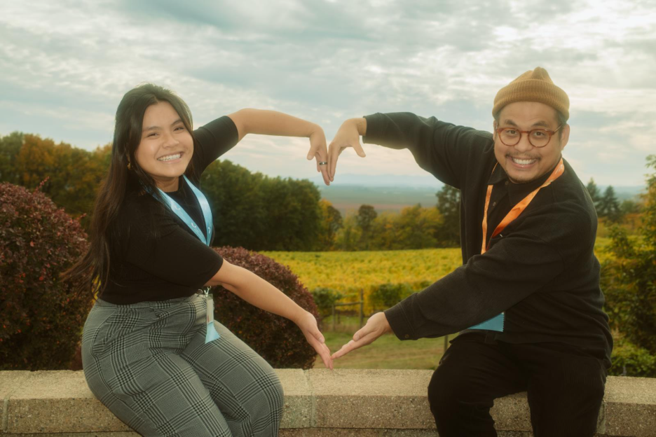 Two people sitting outdoors on a stone ledge with a scenic background of green fields, trees, and a cloudy sky. They are forming a heart shape with their arms and hands.