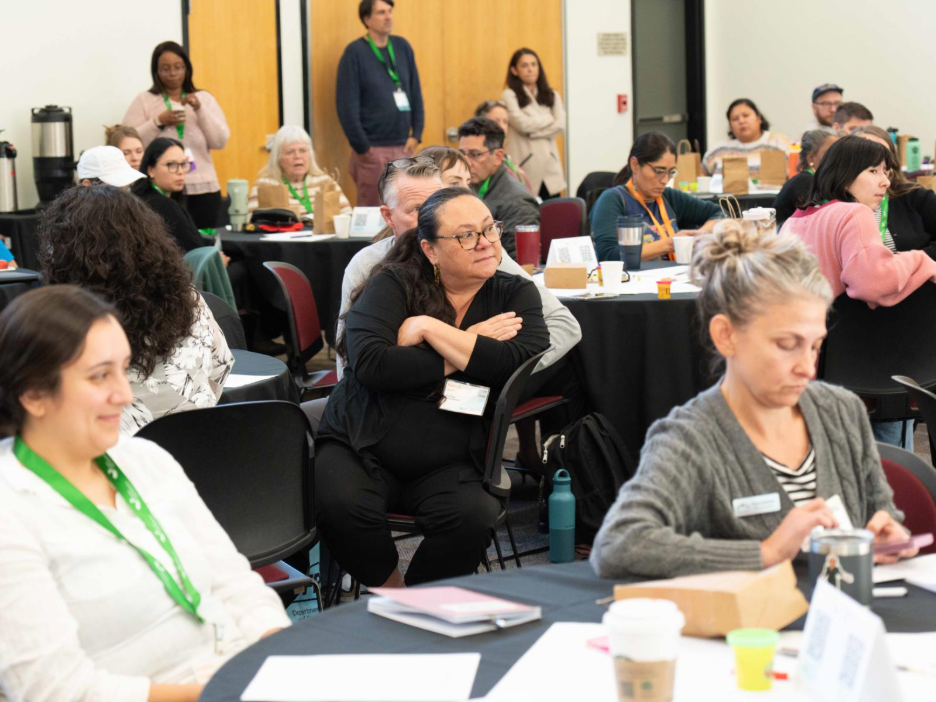 A group of people seated at round tables in a conference room, with notebooks, coffee cups, and name tags visible on the tables. Some individuals are standing near the back, and large beverage dispensers are placed against the wall.
