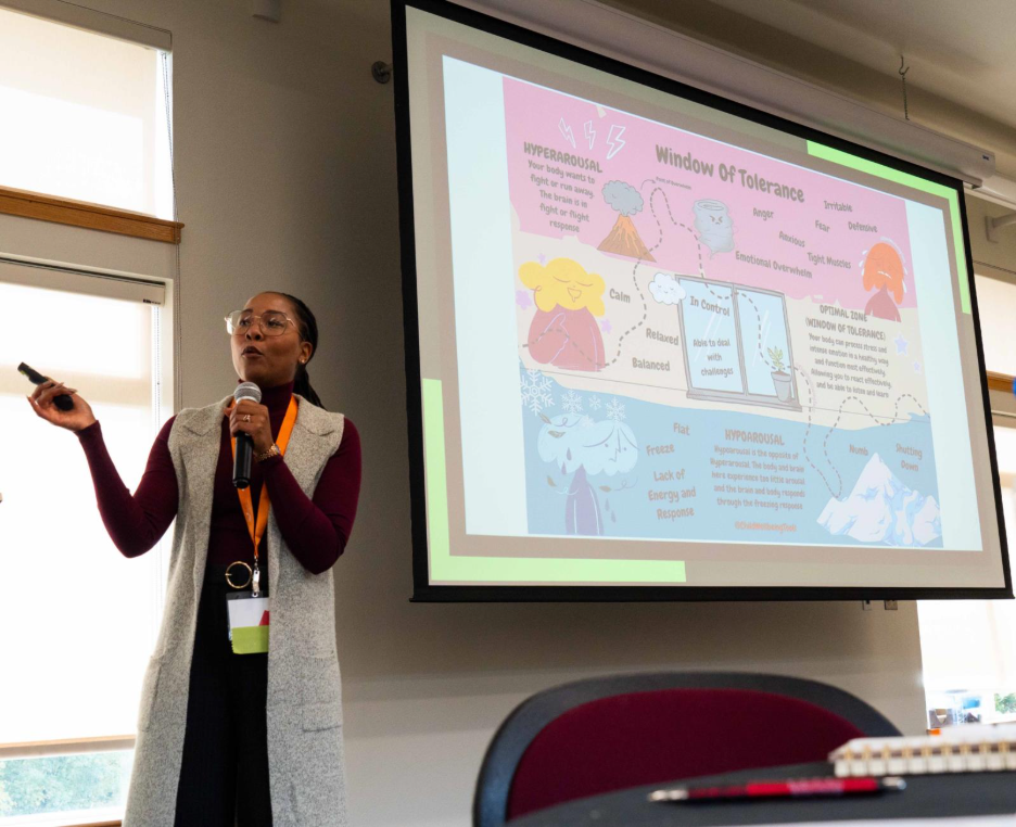 A person standing and presenting in front of a projection screen displaying a colorful diagram titled “Window of Tolerance.” The diagram includes sections labeled hyperarousal, hypoarousal, and optimal zone, with illustrations and descriptive text. A notebook and pen are visible on a table in the foreground.
