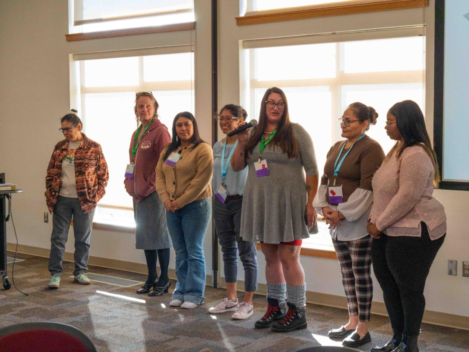 Seven individuals standing in a line inside a well-lit room with large windows, wearing conference badges and casual attire. One person is holding a microphone, suggesting they are speaking or presenting to the group.