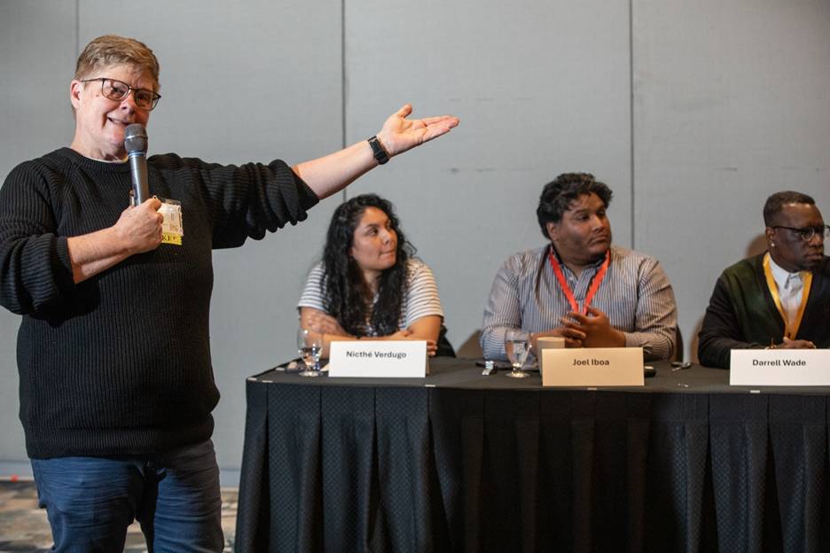 A conference panel discussion with one person standing and speaking into a microphone while gesturing, and three panelists seated at a table listening.